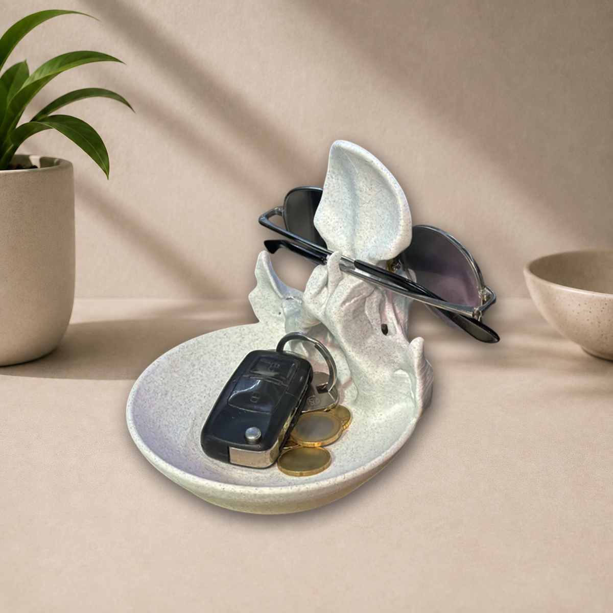 A sculptural ceramic bunny-shaped key holder and sunglasses stand in a styled tabletop scene. The white speckled ceramic piece holds keys and coins in its bowl and supports a pair of sunglasses on its ears.