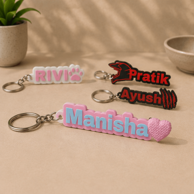 Four custom name keychains on a beige surface, with the blue and pink 'Manisha' keychain and its heart charm in the foreground alongside 'RIVI', 'Pratik', and 'Ayush' keychains.