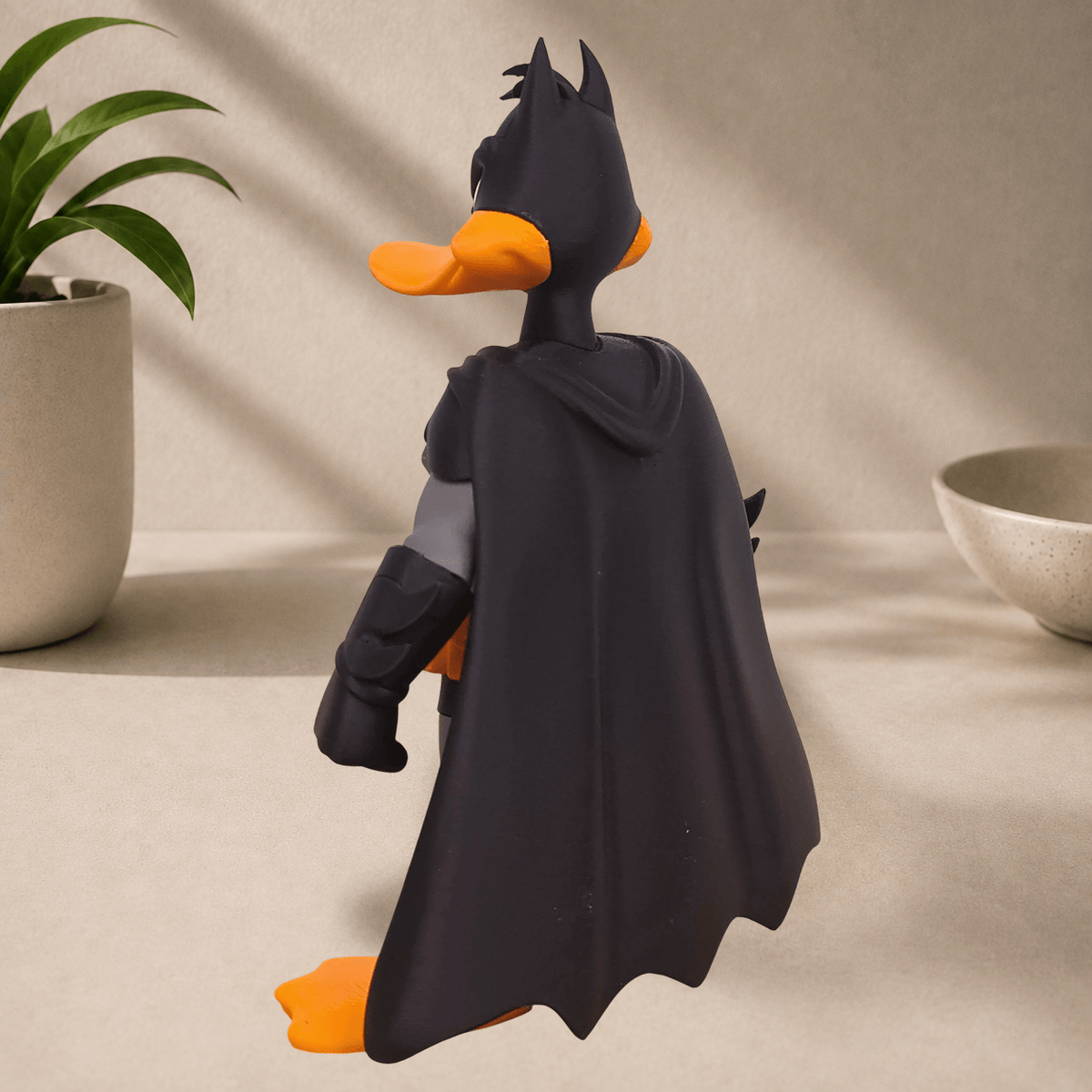 Rear three-quarter view of a Daffy Duck dressed as Batman statue, showing the flowing black cape and pointed-eared cowl, on a beige surface.