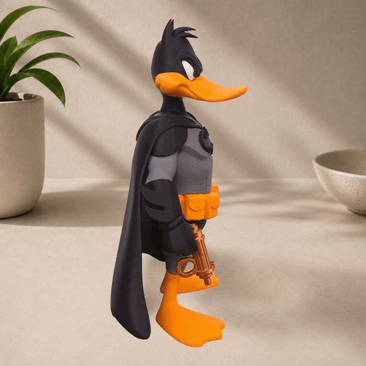 Side profile view of a Daffy Duck dressed as Batman statue in a black cape and gray suit, holding a grappling gun, on a beige surface.