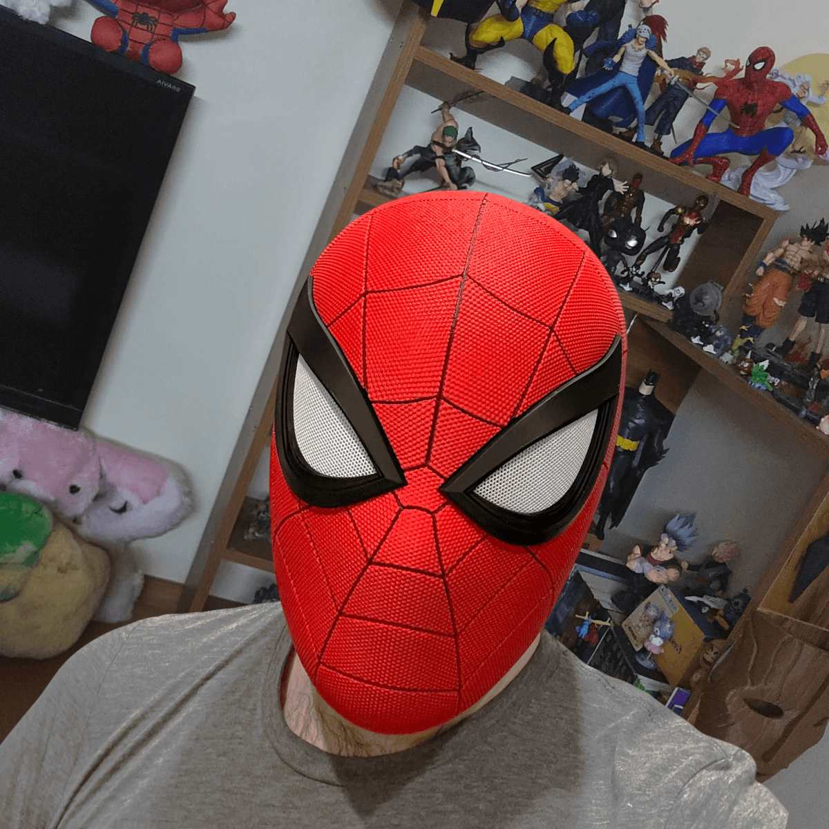 Person wearing the red 3D-printed Spider-Man PS5 cosplay mask, photographed in front of a shelf of collectible figures.