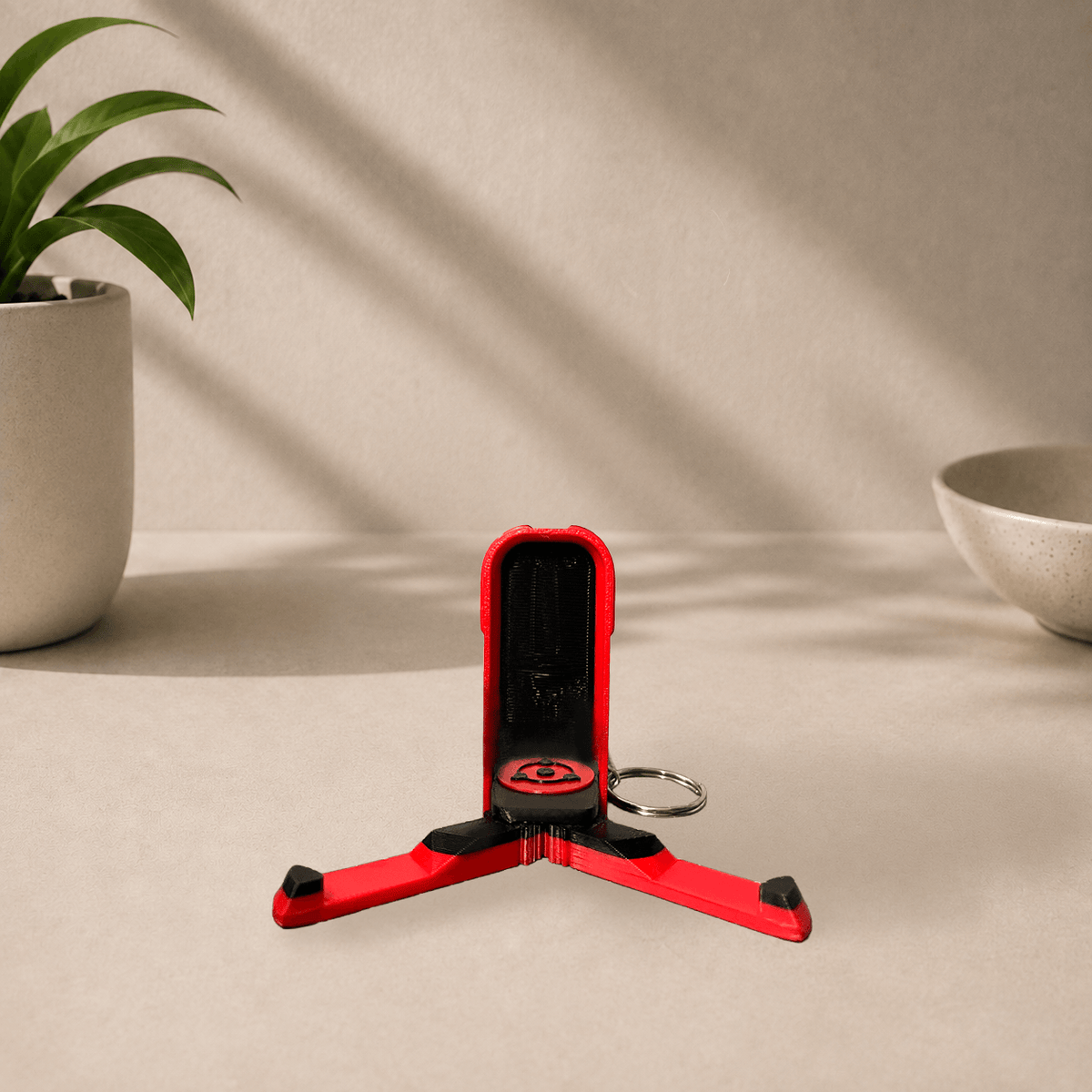 3D-printed Uchiha clan phone stand keychain from Naruto, unfolded into a tripod stand with the red Sharingan emblem facing forward.
