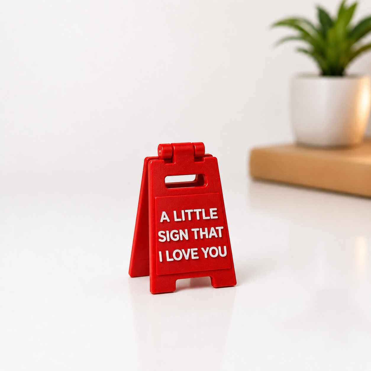 A small red plastic A-frame sign with white text reading 'A LITTLE SIGN THAT I LOVE YOU' on a white surface, with a blurred potted plant in the background.