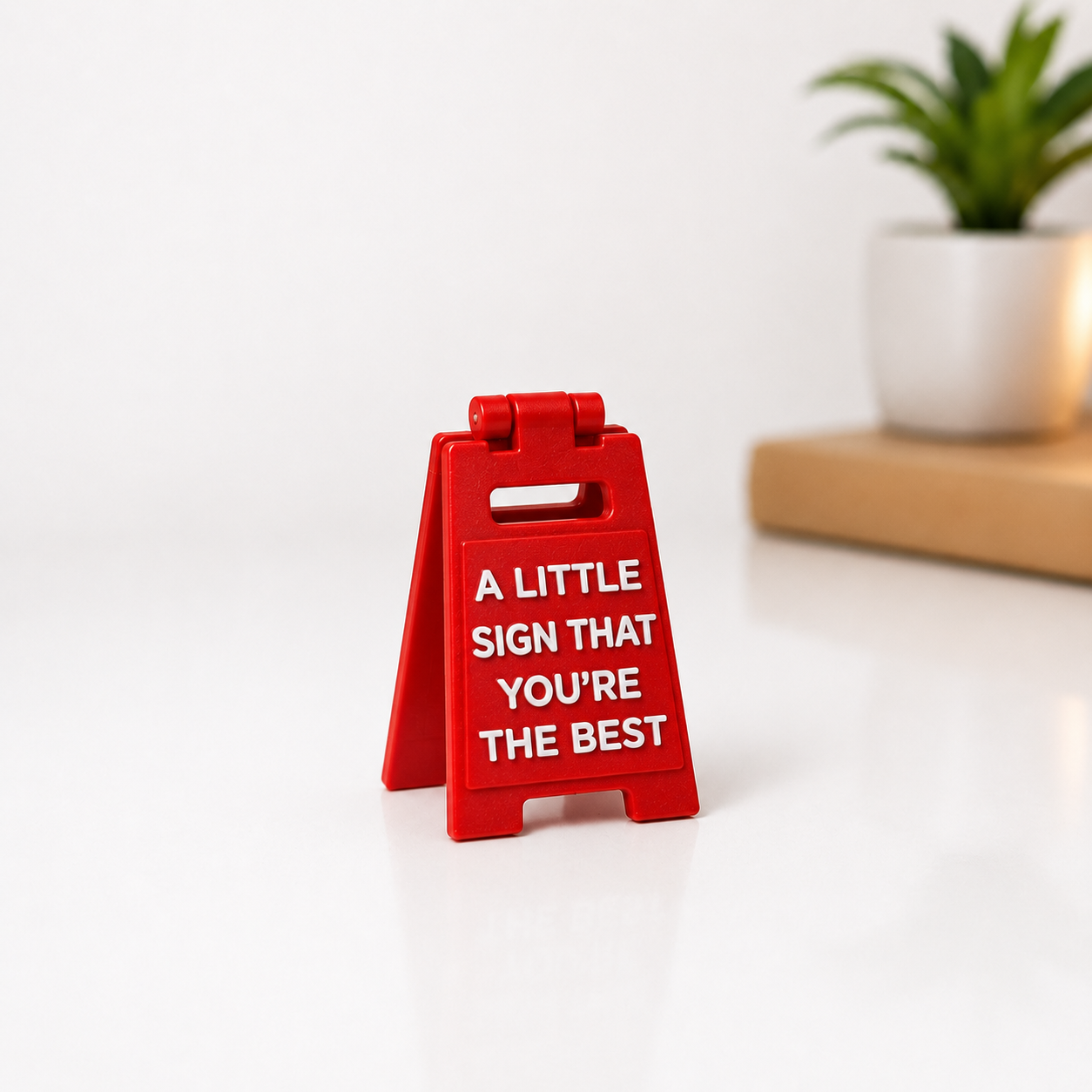 A small red A-frame sign with white text reading 'A LITTLE SIGN THAT YOU'RE THE BEST' on a white surface, with a blurred potted plant in the background.