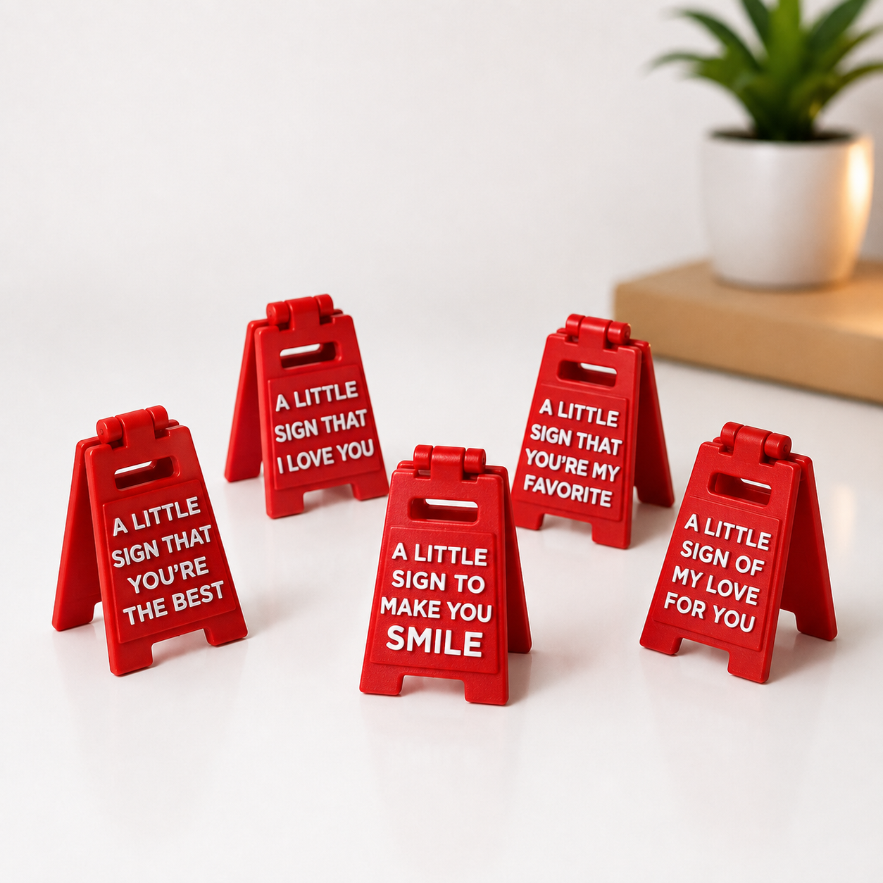 Five small red A-frame sign stands with white text, arranged on a white surface. The signs read phrases such as 'A LITTLE SIGN THAT YOU'RE THE BEST,' 'A LITTLE SIGN THAT I LOVE YOU,' 'A LITTLE SIGN THAT YOU'RE MY FAVORITE,' 'A LITTLE SIGN TO MAKE YOU SMILE,' and 'A LITTLE SIGN OF MY LOVE FOR YOU.