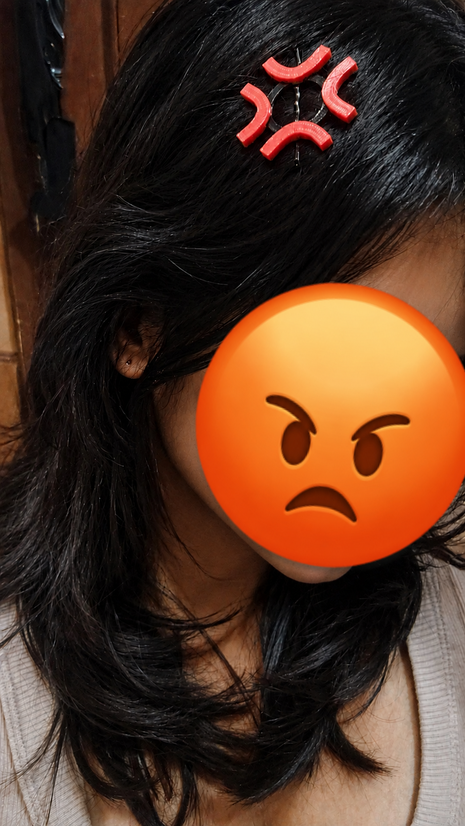 A close-up of a person with long black hair wearing a red angry face hair clip, with an orange angry emoji overlay covering the face.