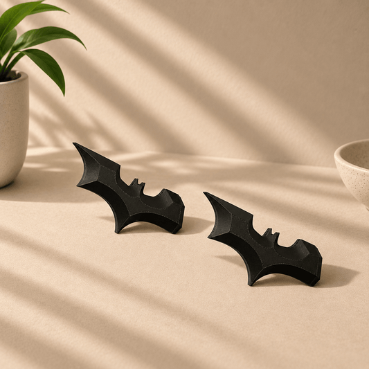 Pair of black 3D-printed Batarang-shaped fridge magnets displayed on a beige tabletop with a potted plant nearby.