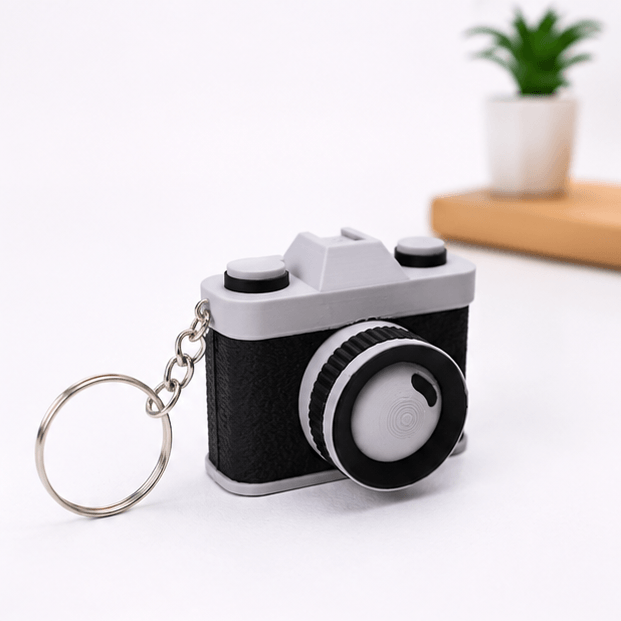 Miniature vintage-camera fidget keychain in black and gray shown at an angled three-quarter view with its silver keyring and chain, on a white background.