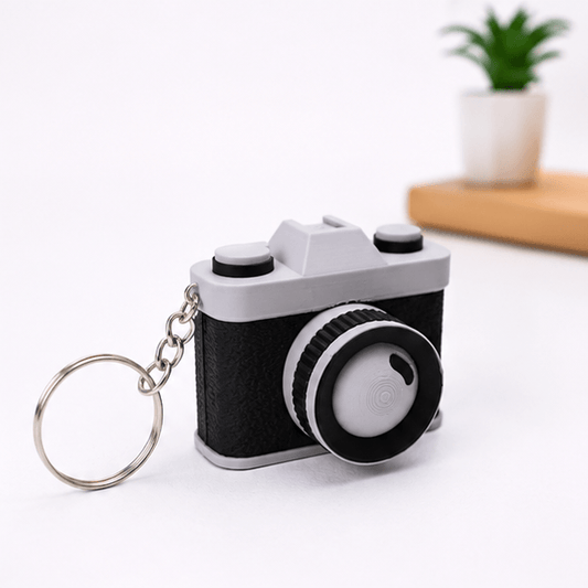 Miniature vintage-camera fidget keychain in black and gray shown at an angled three-quarter view with its silver keyring and chain, on a white background.