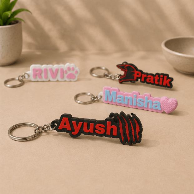 Four custom name keychains on a beige surface, with the red 'Ayush' claw-mark keychain in the foreground alongside 'RIVI', 'Pratik', and 'Manisha' keychains.