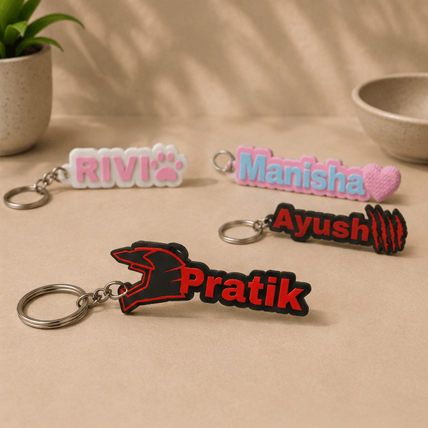 Four custom name keychains on a beige surface, with the red 'Pratik' helmet-design keychain in the foreground alongside 'RIVI', 'Manisha', and 'Ayush' keychains.