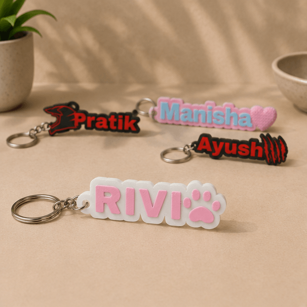 Four custom name keychains on a beige surface, with the pink 'RIVI' paw-print keychain in the foreground alongside 'Pratik', 'Manisha', and 'Ayush' keychains.