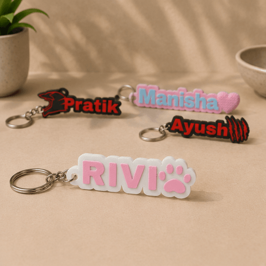 Four custom name keychains on a beige surface, with the pink 'RIVI' paw-print keychain in the foreground alongside 'Pratik', 'Manisha', and 'Ayush' keychains.