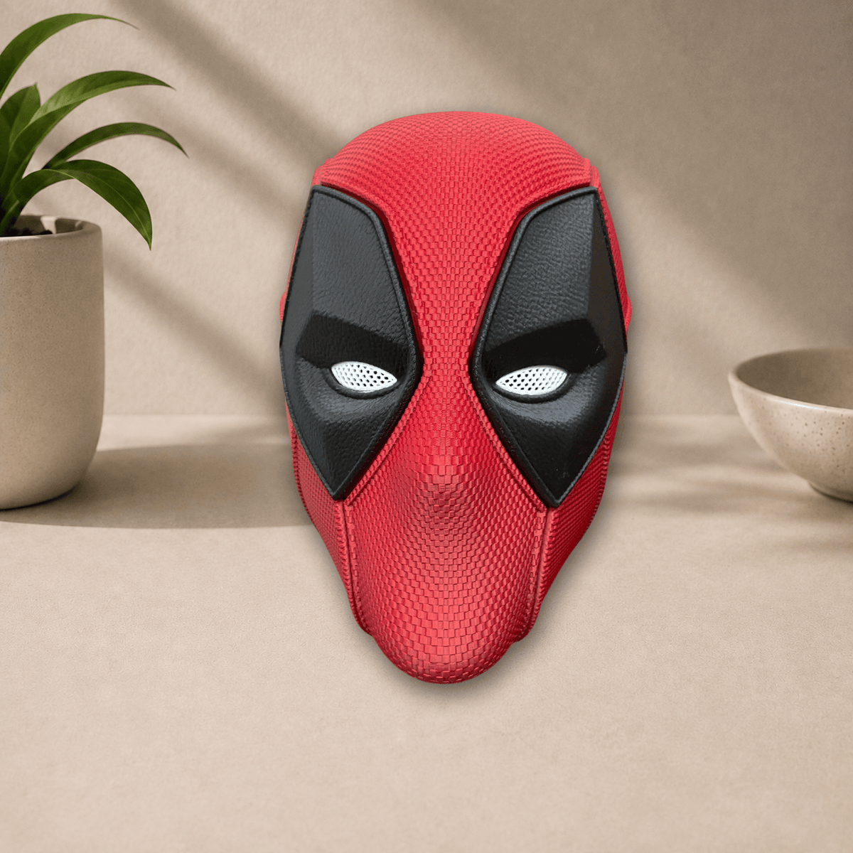 Red and black 3D-printed Deadpool cosplay mask with diamond-shaped eye lenses, front view on a beige tabletop.