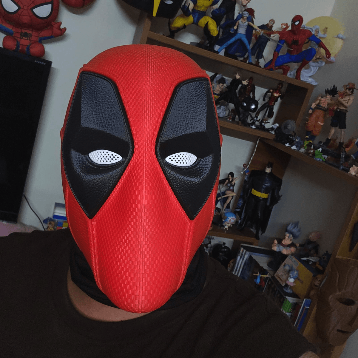 Person wearing the red and black 3D-printed Deadpool cosplay mask in front of a shelf of collectible figures.