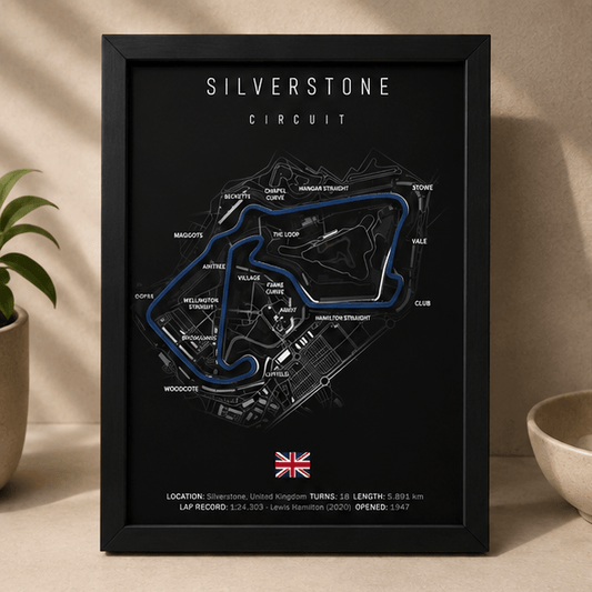 Black-framed 3D-printed Silverstone Circuit F1 track print with a blue highlighted route, standing upright on a table beside potted plants.