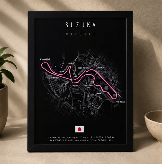 Black-framed 3D-printed Suzuka Circuit F1 track print with a pink highlighted route, standing upright on a table beside a potted plant.