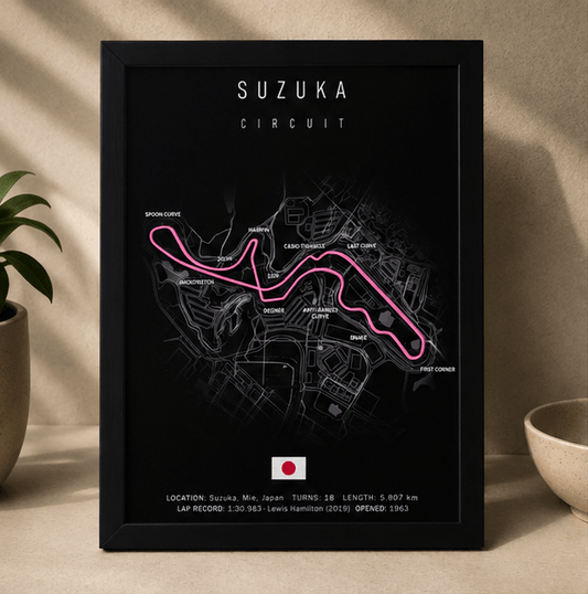 Black-framed 3D-printed Suzuka Circuit F1 track print with a pink highlighted route, standing upright on a table beside a potted plant.