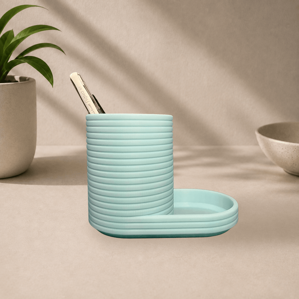 Mint green 3D-printed ribbed Flow pen holder with an attached oval tray, holding a pen, on a beige tabletop.