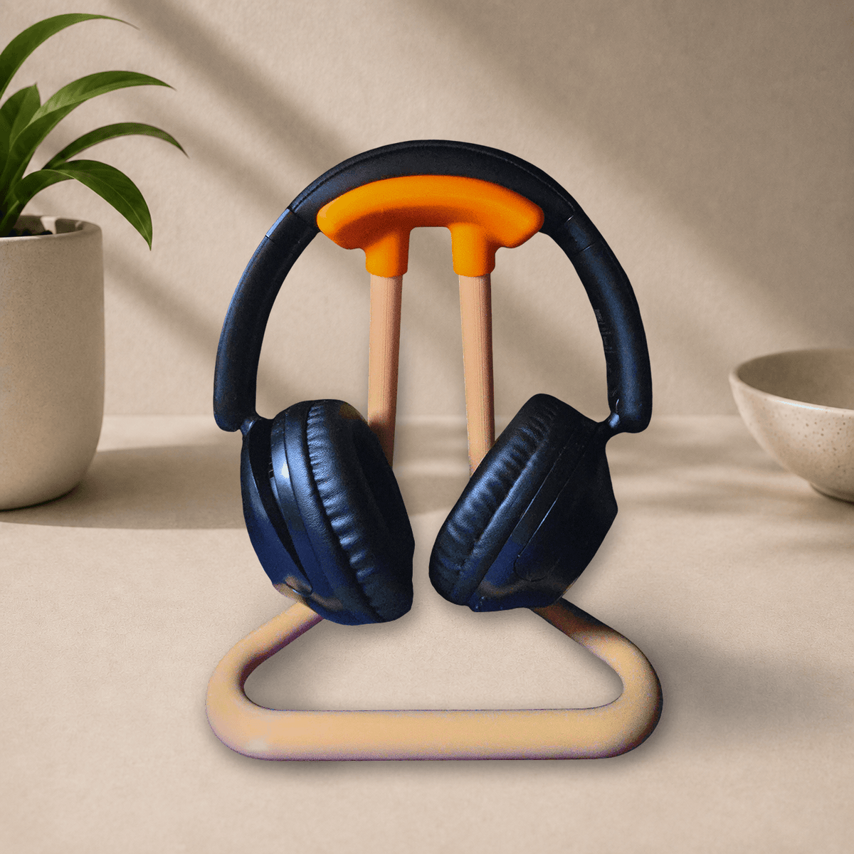 Black headphones resting on a 3D-printed desk stand with orange grip and tan legs, beige desk background with a plant and bowl