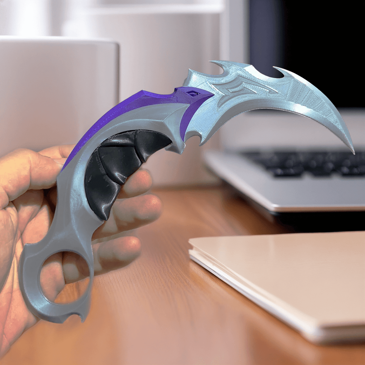 Hand holding the 3D-printed Reaver Karambit display prop next to a laptop and notebook on a wooden desk