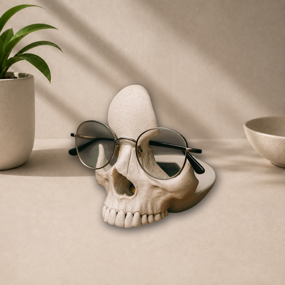 White 3D-printed skull-shaped glasses holder with a pair of round-frame glasses resting on its nose bridge, displayed on a beige tabletop.