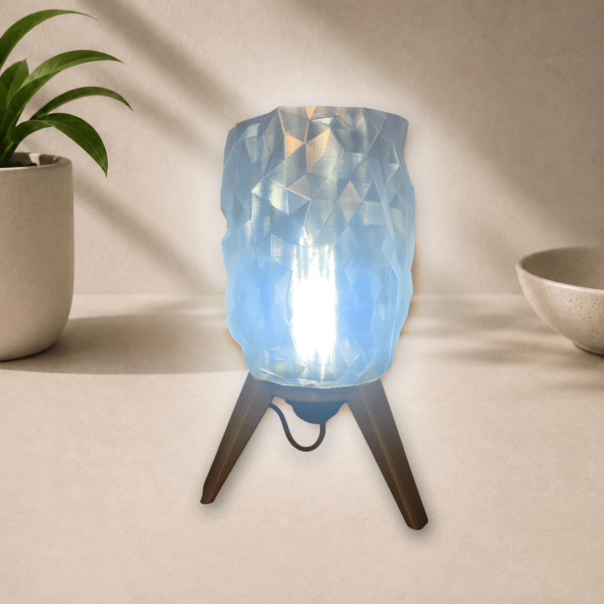 3D-printed tripod table lamp with a diamond-faceted shade on gold-toned legs, glowing cool blue-white when lit.