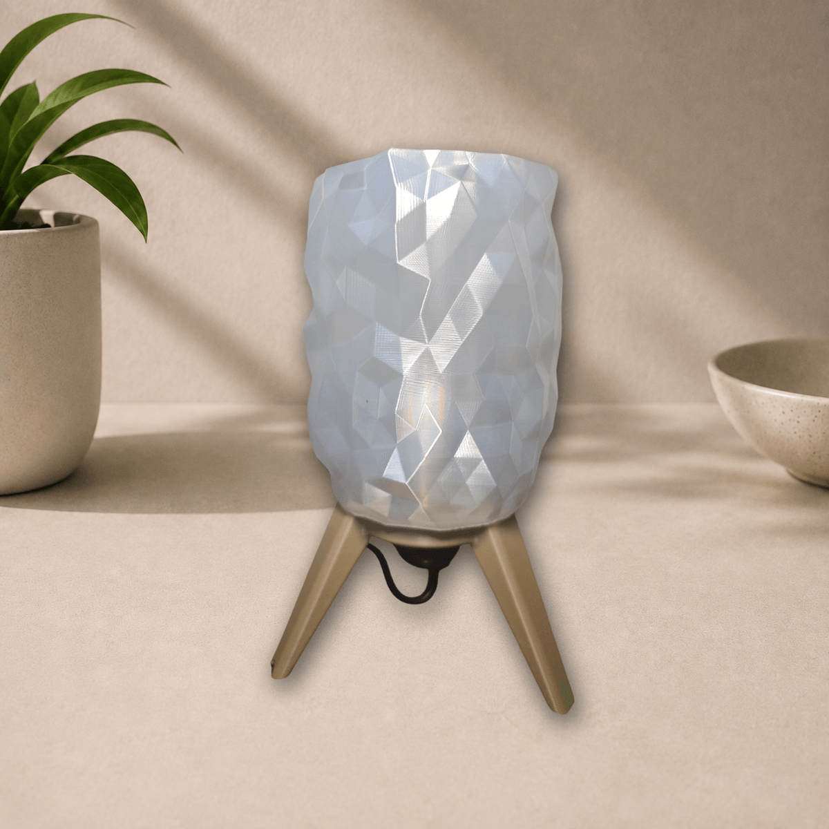 3D-printed tripod table lamp with a diamond-faceted white shade on gold-toned tripod legs, unlit, front view on a beige tabletop.