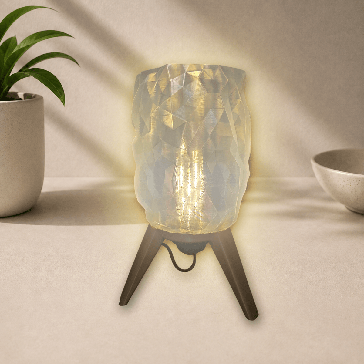 3D-printed tripod table lamp with a diamond-faceted shade on gold-toned legs, glowing warm amber when lit.