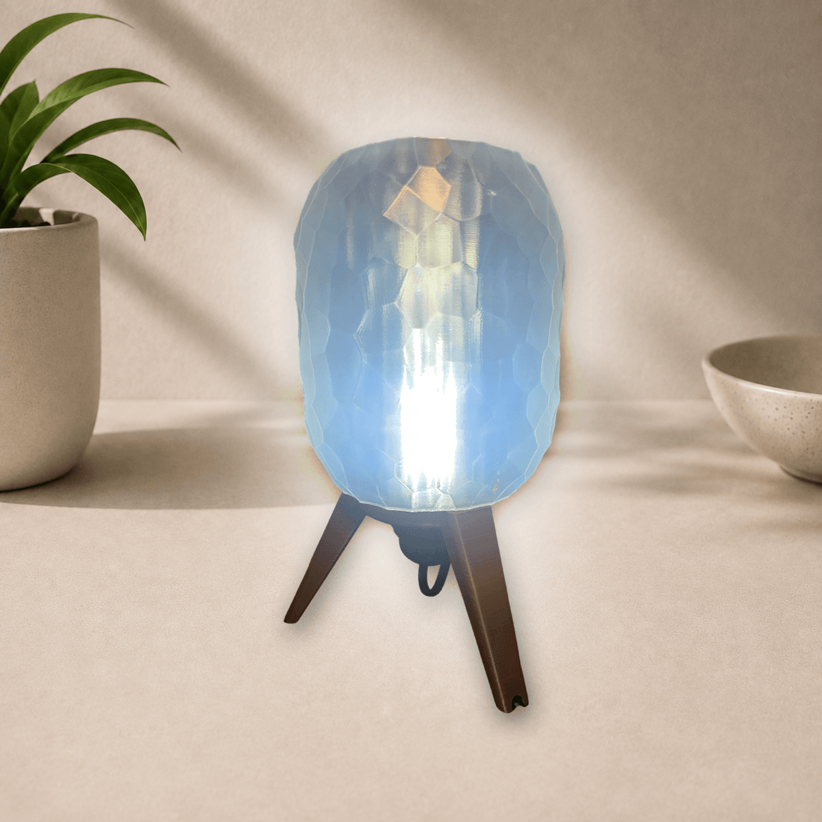 3D-printed tripod table lamp with a pebble-textured shade on dark bronze legs, glowing cool blue-white when lit.