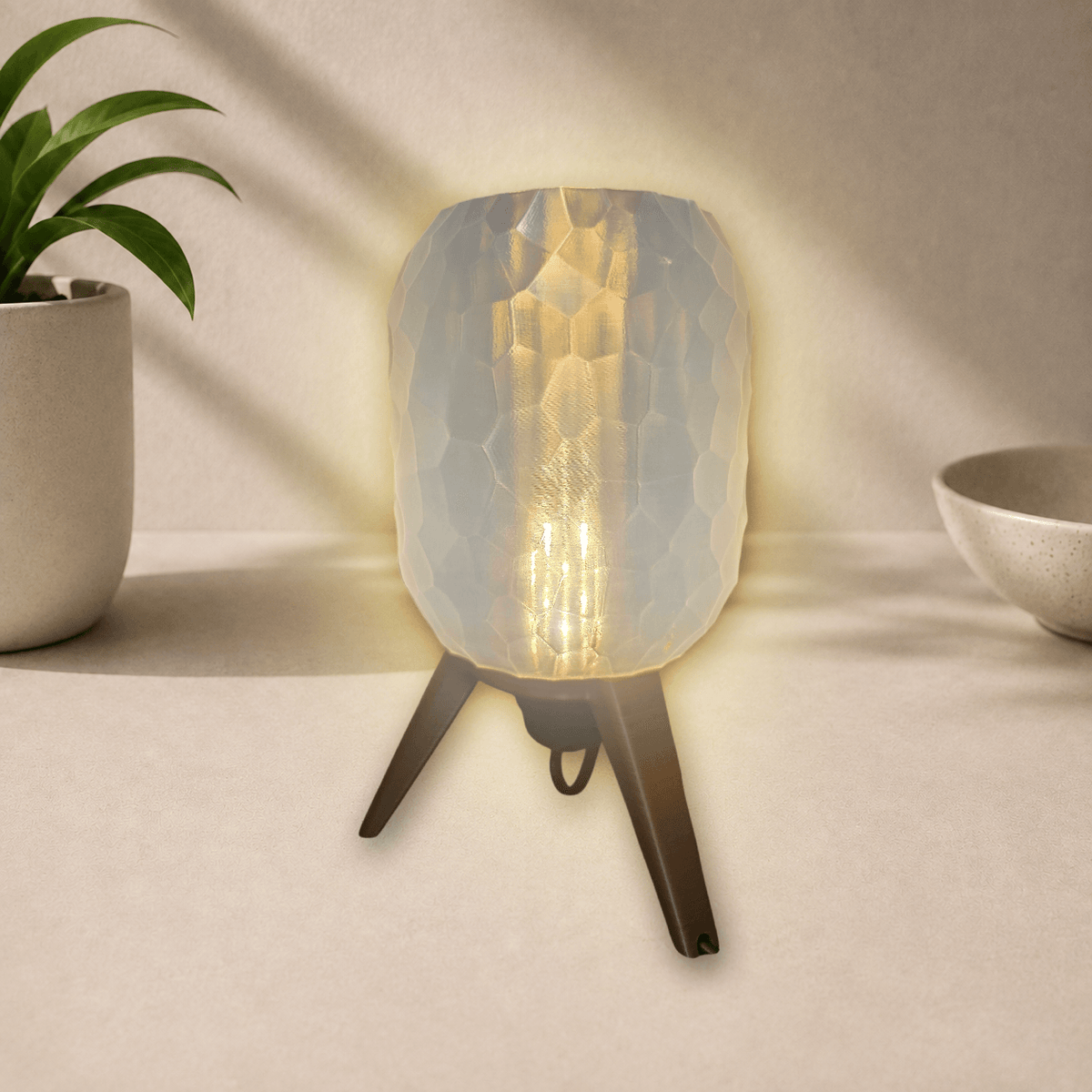 3D-printed tripod table lamp with a pebble-textured shade on dark bronze legs, glowing warm amber when lit.